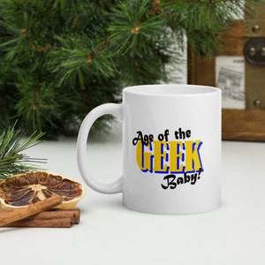 Könnte beinhalten: Weiße Keramiktasse mit einer schwarzen und blauen Grafik, auf der "Age of the Geek Baby!" steht. Der Text befindet sich in einem gelben Kasten mit blauem Rand.