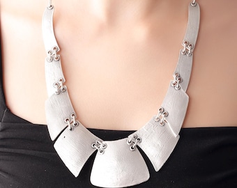 May include: A silver statement necklace with a geometric design. The necklace is made of multiple connected pieces that form a wide, curved shape.
