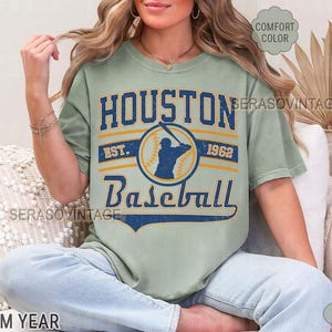 Puede incluir: Una camiseta verde claro con un dise&ntilde;o de b&eacute;isbol vintage. La camiseta tiene el texto "Houston Baseball" con los a&ntilde;os "Est. 1962".