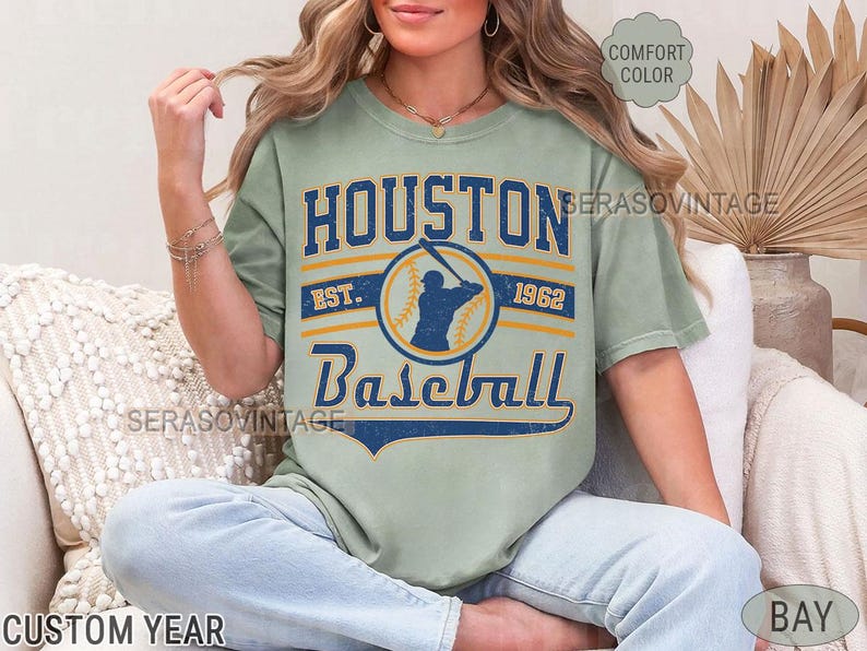 Puede incluir: Una camiseta verde claro con un dise&ntilde;o de b&eacute;isbol vintage. La camiseta tiene el texto "Houston Baseball" con los a&ntilde;os "Est. 1962".