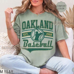 May include: A green t-shirt with a vintage Oakland Baseball logo. The logo features a baseball player with a bat and the text "Oakland Baseball Est. 1968".