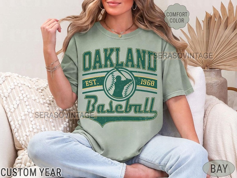 May include: A green t-shirt with a vintage Oakland Baseball logo. The logo features a baseball player with a bat and the text "Oakland Baseball Est. 1968".