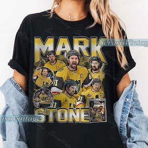 以下が含まれることがあります： 黒いTシャツ。金と黒のユニフォームを着たホッケー選手、マーク・ストーンのグラフィックが特徴です。「MARK STONE」の文字が金色で、選手の複数の画像が描かれています。ヴィンテージ感のあるデザインです。