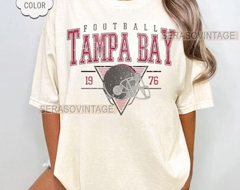 Puede incluir: Camiseta de color marfil con un diseño de fútbol vintage. El diseño presenta las palabras "TAMPA BAY" en letras rojas, un casco de fútbol y los números "19" y "76". La camiseta tiene una etiqueta "Comfort Color".
