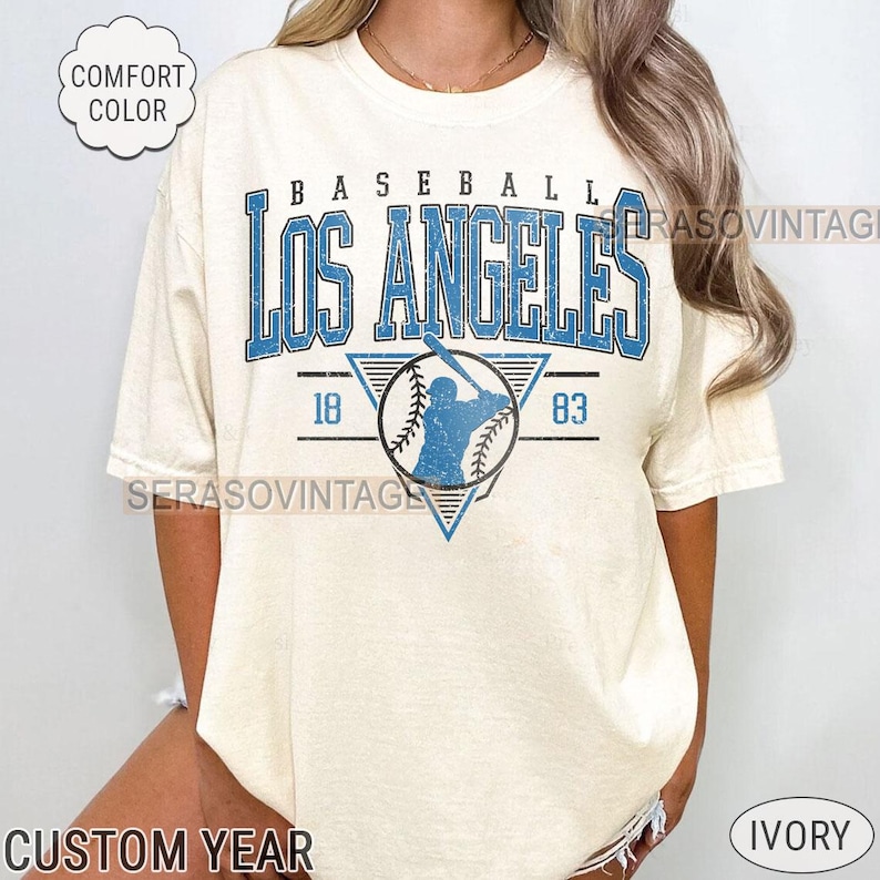 以下が含まれることがあります： クリーム色のTシャツに、青と白で「Baseball Los Angeles」の文字が書かれています。Tシャツには、青と白の野球選手のグラフィックも描かれており、両側に「18」と「83」の数字があります。