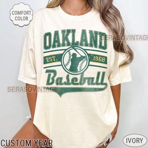 May include: A white t-shirt with a green and gold graphic that says "Oakland Baseball Est. 1968". The graphic features a baseball player in a silhouette.