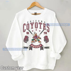 Puede incluir: Sudadera blanca con un logotipo vintage de hockey de los Arizona Coyotes. El logotipo presenta un coyote mascota sosteniendo un palo de hockey y vistiendo una camiseta roja y negra. El texto "Arizona Coyotes" est&aacute; en rojo y negro con un aspecto desgastado. El texto "Hockey" est&aacute; en negro con un aspecto desgastado. Los n&uacute;meros "19" y "72" est&aacute;n a cada lado del logotipo.