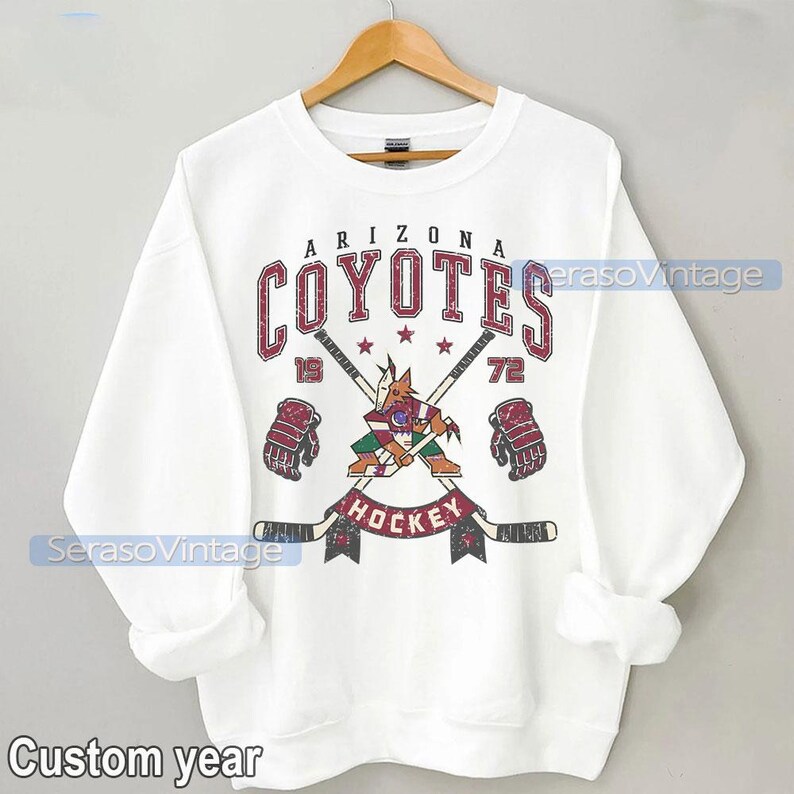 Puede incluir: Sudadera blanca con un logotipo vintage de hockey de los Arizona Coyotes. El logotipo presenta un coyote mascota sosteniendo un palo de hockey y vistiendo una camiseta roja y negra. El texto "Arizona Coyotes" est&aacute; en rojo y negro con un aspecto desgastado. El texto "Hockey" est&aacute; en negro con un aspecto desgastado. Los n&uacute;meros "19" y "72" est&aacute;n a cada lado del logotipo.