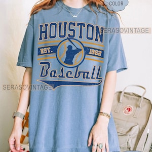 Puede incluir: Una camiseta azul con un gr&aacute;fico de estilo vintage de un jugador de b&eacute;isbol y el texto "Houston Baseball Est. 1962".