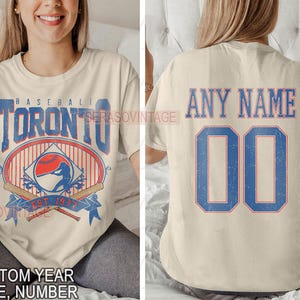 May include: Two cream-coloured t-shirts with a baseball theme. The front shirt has a vintage Toronto baseball logo with a baseball player and the text "Baseball Toronto Est 1977". The back shirt has the text "Any Name" and a large number "00" in blue with a red outline.