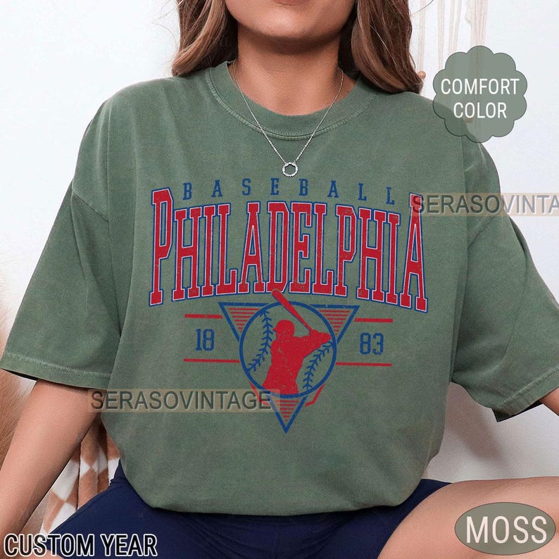 Puede incluir: Una camiseta verde con un gr&aacute;fico de b&eacute;isbol vintage. El gr&aacute;fico presenta un jugador de b&eacute;isbol rojo con un bate, una pelota de b&eacute;isbol y el texto "BASEBALL PHILADELPHIA 18 83".