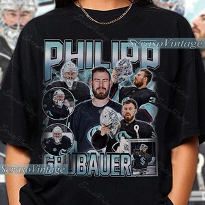 Puede incluir: Camiseta negra con un diseño en verde azulado y blanco que presenta a Philipp Grubauer, un portero de hockey. El diseño incluye el nombre "PHILIPP GRUBAUER" y varias imágenes del jugador y su máscara de portero.