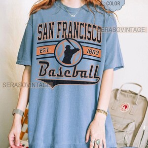 May include: A blue t-shirt with a vintage baseball graphic. The graphic features a baseball player in silhouette, the text "San Francisco Baseball", and the years "Est. 1883".
