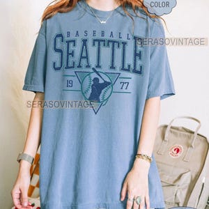 May include: A blue t-shirt with a vintage baseball graphic. The graphic features a baseball player in a triangle with the text "BASEBALL SEATTLE 19 77" above the player. The shirt is a comfortable fit and is available in a variety of colors.
