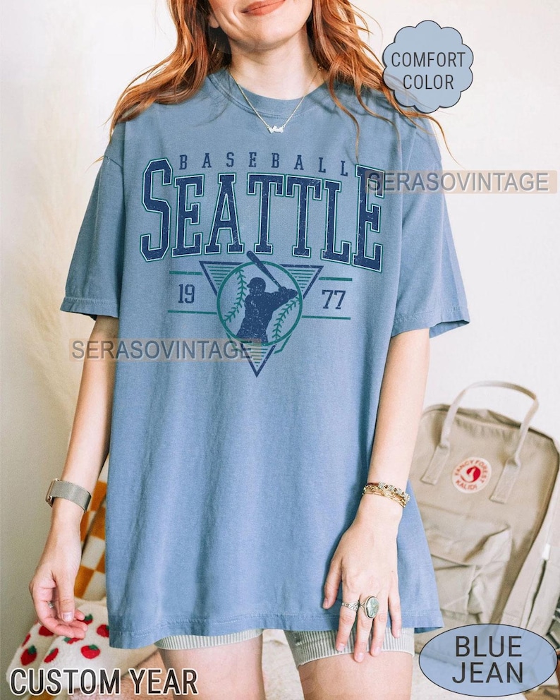 May include: A blue t-shirt with a vintage baseball graphic. The graphic features a baseball player in a triangle with the text "BASEBALL SEATTLE 19 77" above the player. The shirt is a comfortable fit and is available in a variety of colors.