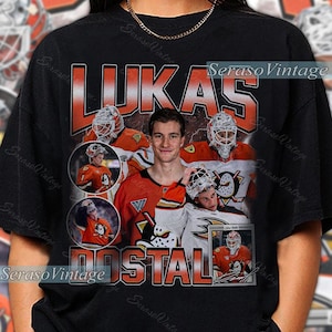 Op de afbeelding: Zwart T-shirt met een ontwerp met een hockeyspeler, met de naam "LUKAS POSTAL" in oranje en witte tekst. Het ontwerp bevat afbeeldingen van de speler in zijn hockey-uniform en met zijn goalie-masker.