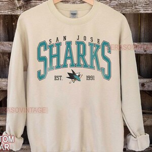 May include: A cream colored sweatshirt with the words "San Jose Sharks" in teal and navy blue. The logo of the San Jose Sharks hockey team is printed below the text. The text "Est. 1991" is printed below the logo.