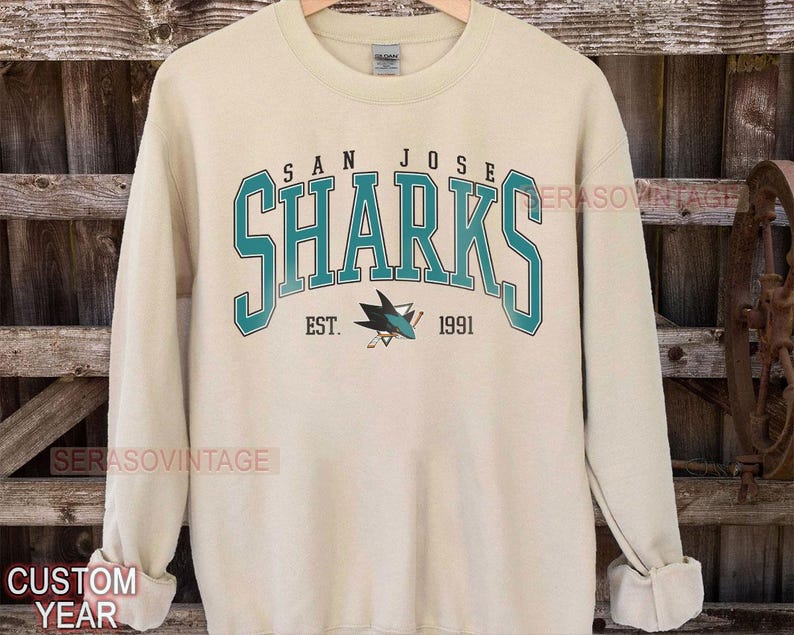 May include: A cream colored sweatshirt with the words "San Jose Sharks" in teal and navy blue. The logo of the San Jose Sharks hockey team is printed below the text. The text "Est. 1991" is printed below the logo.