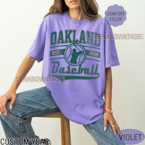 May include: A purple t-shirt with a vintage-style graphic of a baseball player and the text "Oakland Baseball Est. 1968".