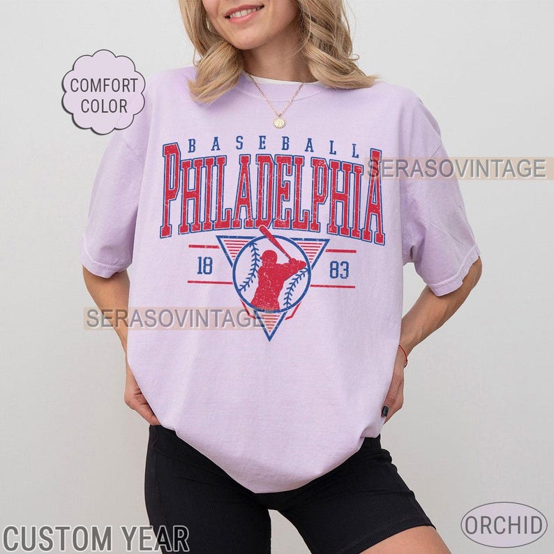 Puede incluir: Una camiseta lavanda con un gr&aacute;fico de b&eacute;isbol vintage. El gr&aacute;fico presenta a un jugador de b&eacute;isbol en rojo y azul, con el texto "BASEBALL PHILADELPHIA 18 83" en azul y rojo.