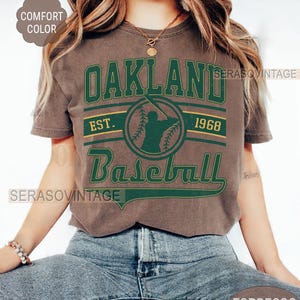 May include: A brown t-shirt with a green and gold graphic that says "Oakland Baseball Est. 1968".