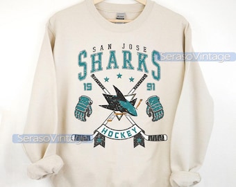 May include: A beige sweatshirt featuring a vintage San Jose Sharks hockey logo. The logo depicts a teal shark, crossed hockey sticks, and the text "San Jose Sharks Hockey".