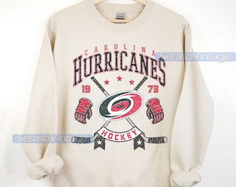 Puede incluir: Una sudadera beige con un logotipo de hockey de los Carolina Hurricanes. El logotipo presenta un símbolo de huracán rojo y negro con palos de hockey cruzados y el texto "Carolina Hurricanes" y "Hockey". Los números "19" y "73" están a cada lado del logotipo.