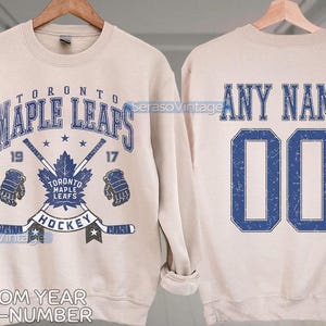 Puede incluir: Una sudadera beige con un logotipo de hockey de los Toronto Maple Leafs azul y blanco en la parte delantera y el texto "TORONTO MAPLE LEAFS HOCKEY" y "19" y "17". La parte posterior de la sudadera tiene el texto "ANY NAME" y "00" en azul.