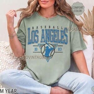 以下が含まれることがあります： ヴィンテージな野球のグラフィックがプリントされた薄い緑色のTシャツ。グラフィックには、青いシルエットの野球選手と「Baseball Los Angeles 18 83」の文字が描かれています。