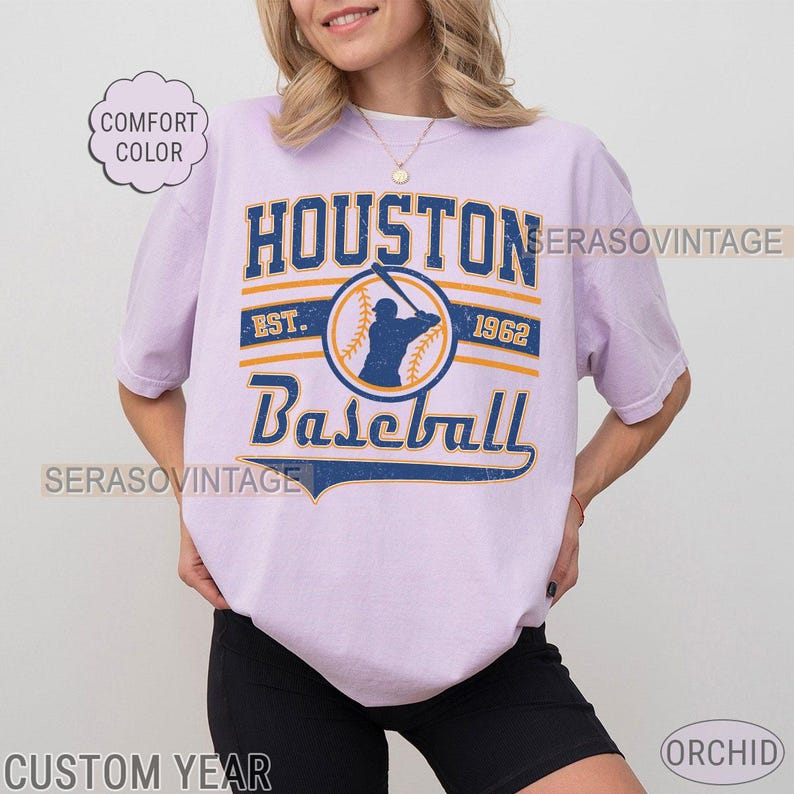 Puede incluir: Una camiseta lavanda con un gr&aacute;fico de b&eacute;isbol vintage. El gr&aacute;fico presenta a un jugador de b&eacute;isbol en un c&iacute;rculo con el texto "Houston Baseball Est. 1962".
