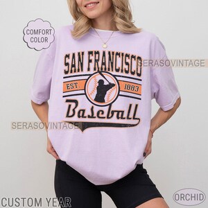 May include: A lavender t-shirt with a vintage baseball graphic. The graphic features the text "San Francisco Baseball" with the years "Est. 1883" and a silhouette of a baseball player.