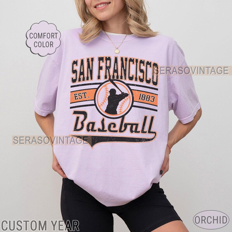 May include: A lavender t-shirt with a vintage baseball graphic. The graphic features the text "San Francisco Baseball" with the years "Est. 1883" and a silhouette of a baseball player.