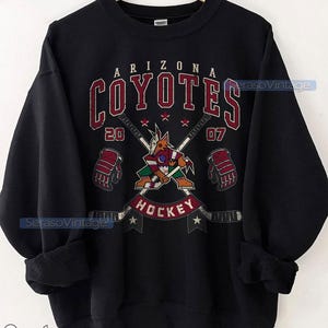 Puede incluir: Sudadera negra con un logotipo de hockey de los Arizona Coyotes en rojo, blanco y negro. El logotipo representa un coyote sosteniendo un palo de hockey con el texto "Arizona Coyotes" y "Hockey". Los n&uacute;meros "20" y "07" est&aacute;n colocados a cada lado del logotipo.