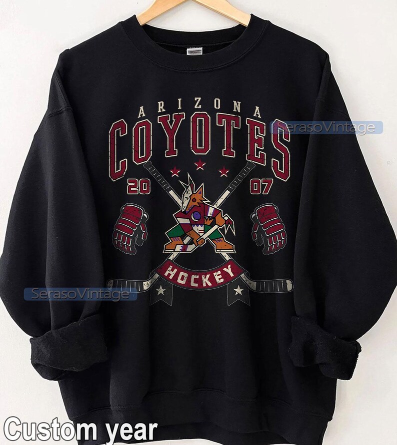 Puede incluir: Sudadera negra con un logotipo de hockey de los Arizona Coyotes en rojo, blanco y negro. El logotipo representa un coyote sosteniendo un palo de hockey con el texto "Arizona Coyotes" y "Hockey". Los n&uacute;meros "20" y "07" est&aacute;n colocados a cada lado del logotipo.