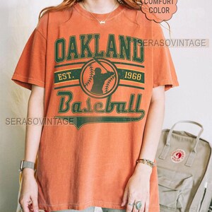 May include: A burnt orange t-shirt with a green and white graphic that says "Oakland Baseball Est. 1968".