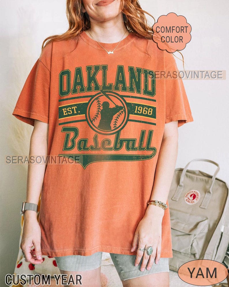 May include: A burnt orange t-shirt with a green and white graphic that says "Oakland Baseball Est. 1968".