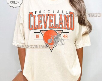 May include: Ivory t-shirt featuring the word "FOOTBALL" above "CLEVELAND" in orange block letters. A football helmet graphic is centered below the text, with the numbers "19" and "46" on either side. The shirt has a "COMFORT COLOR" label.
