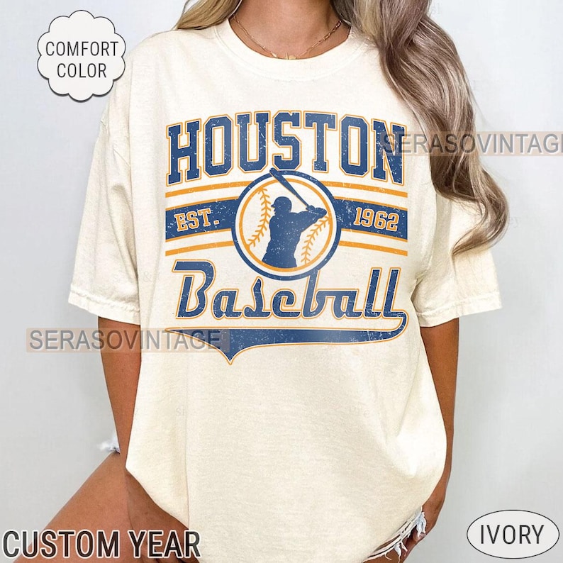 Puede incluir: Una camiseta de color crema con un dise&ntilde;o gr&aacute;fico vintage que presenta el texto "Houston Baseball" con una silueta de jugador de b&eacute;isbol y el texto "Est. 1962".