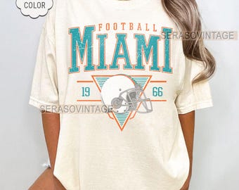 Könnte beinhalten: Cremefarbenes T-Shirt mit dem Wort "FOOTBALL" über "MIAMI" in türkisfarbenen Blockbuchstaben. Darunter befindet sich eine Footballhelm-Grafik, die in einem Dreieck zentriert ist, mit den Zahlen "19" und "66" auf beiden Seiten. Das Shirt hat ein "Comfort Color"-Label.