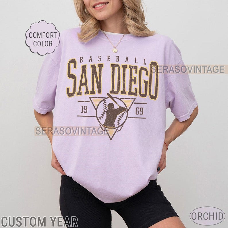 Może przedstawiać: Fioletowy t-shirt z vintage grafiką baseballową. Grafika przedstawia gracza baseballowego w tr&oacute;jkącie z tekstem "BASEBALL SAN DIEGO 19 69" w kolorze ż&oacute;łtym.