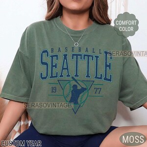 May include: A green t-shirt with a vintage baseball design. The design features the word "SEATTLE" in a blue and white font, a baseball player silhouette, and the numbers "19" and "77".