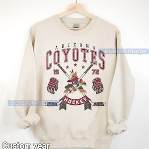 Puede incluir: Una sudadera beige con un logotipo vintage de hockey de los Arizona Coyotes. El logotipo presenta un coyote mascota sosteniendo un palo de hockey con palos cruzados y las palabras "Arizona Coyotes" y "Hockey" en una fuente desgastada.