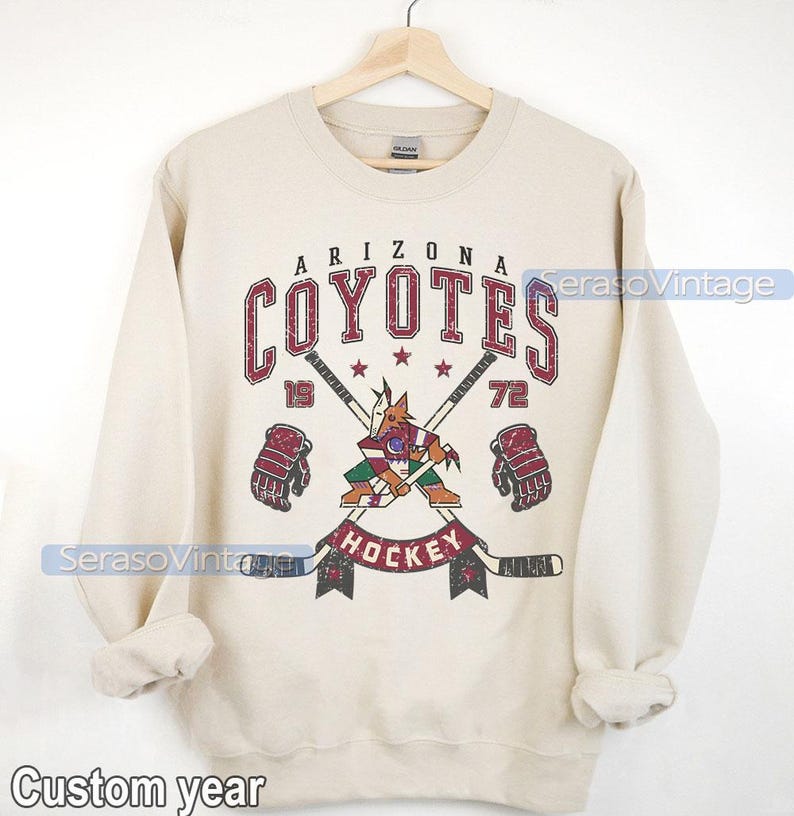 Puede incluir: Una sudadera beige con un logotipo vintage de hockey de los Arizona Coyotes. El logotipo presenta un coyote mascota sosteniendo un palo de hockey con palos cruzados y las palabras "Arizona Coyotes" y "Hockey" en una fuente desgastada.
