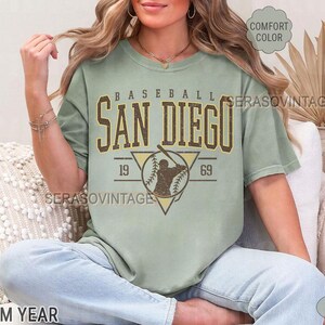 Może przedstawiać: Jasnozielony t-shirt z vintage grafiką baseballową. Grafika przedstawia baseballistę z tekstem "Baseball San Diego 19 69".