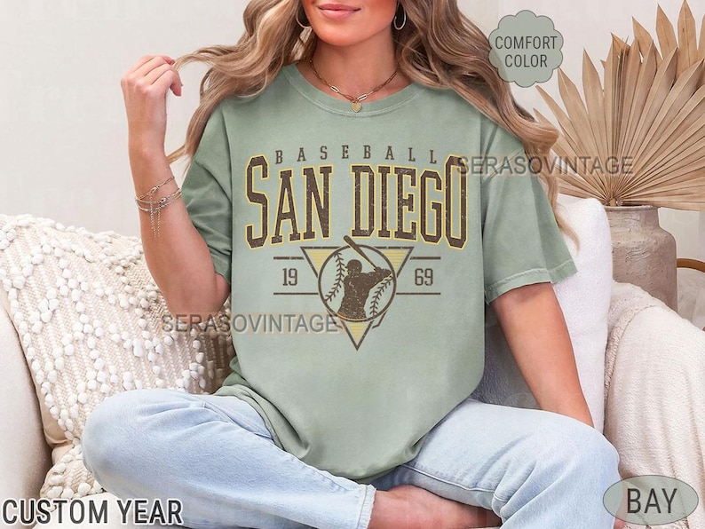 Może przedstawiać: Jasnozielony t-shirt z vintage grafiką baseballową. Grafika przedstawia baseballistę z tekstem "Baseball San Diego 19 69".