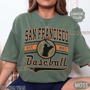 May include: A green t-shirt with a vintage-style graphic design featuring the text "San Francisco Baseball Est. 1883" with a baseball player silhouette and a baseball inside a circle.
