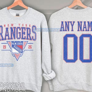 Op de afbeelding: Grijs sweatshirt met een New York Rangers logo op de voorkant en "ANY NAME 00" op de achterkant. Het sweatshirt is aanpasbaar met een jaar, naam en nummer.