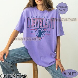 Puede incluir: Una camiseta morada con un gr&aacute;fico de b&eacute;isbol vintage que dice "Cleveland" con los a&ntilde;os "18" y "94" a cada lado del gr&aacute;fico. El gr&aacute;fico es en azul, rojo y blanco. La camiseta es de color confort.