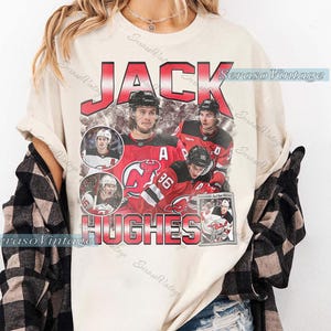 May include: Cream-colored t-shirt featuring a vintage-style graphic of hockey player Jack Hughes. The design includes the name "JACK HUGHES" in red, with images of the player in action. The shirt has a relaxed fit and short sleeves.