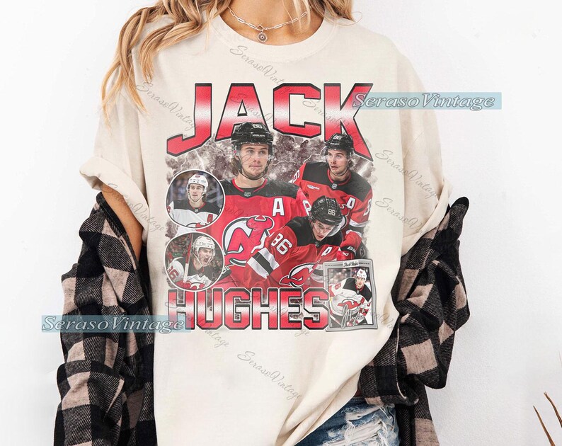 May include: Cream-colored t-shirt featuring a vintage-style graphic of hockey player Jack Hughes. The design includes the name "JACK HUGHES" in red, with images of the player in action. The shirt has a relaxed fit and short sleeves.